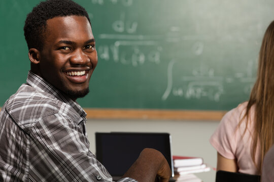 Smiling african american male student looking back in classroom during math lecture with formulas on chalkboard