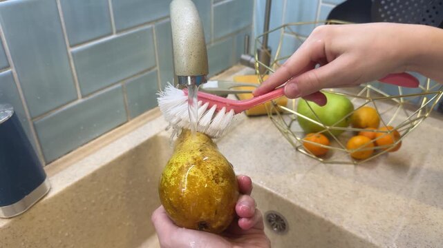 In a sunny kitchen at home, a child who follows a vegan lifestyle gently washes dirt off a freshly picked pear under running water, practicing good hygiene.