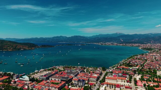 Aerial tracking pan of coastal town harbor with boats and mountainous backdrop over azure bay Fethiye, Turkey.