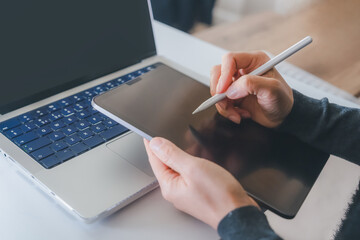 Person using digital stylus pen on tablet beside laptop computer in modern workspace. Remote work, online learning, business technology, creative workflow, and digital communication concept  © Anchalee