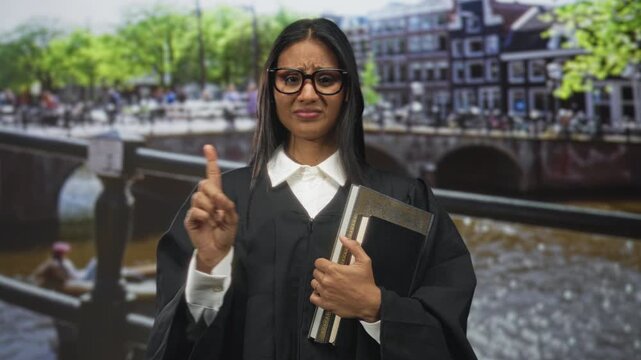 Woman judge brunette wearing robe and glasses holding book and ringed hand points finger in street canal bridge setting; authority concern.