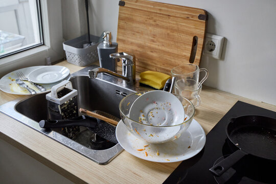 A messy kitchen sink counter filled with dirty dishes, unwashed glass mugs, greasy plates with cutlery, and a cutting board. Domestic chores concept.