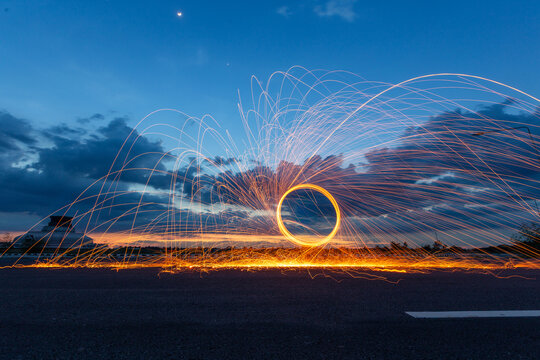 Spinning burning steel wool creates glowing spark trails in a circle at twilight with a temple stupa in the background.
