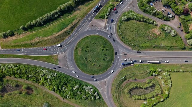 Aerial view of a busy traffic roundabout with cars moving through the junction surrounded by green fields and trees in Kettering, England, United Kingdom.