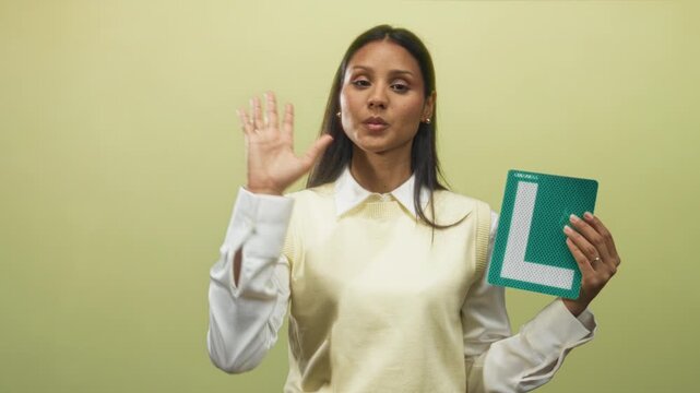 Woman holds green learner l plate with right hand and gestures with left hand while demonstrating driving steps in studio; driver education confidence.
