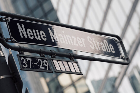 Frankfurt street signage for Neue Mainzer Strasse sits before blurred office glass, tying facade lines and district architecture to place