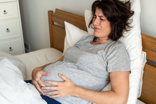 Pregnant woman in discomfort lying in bed, holding her belly.