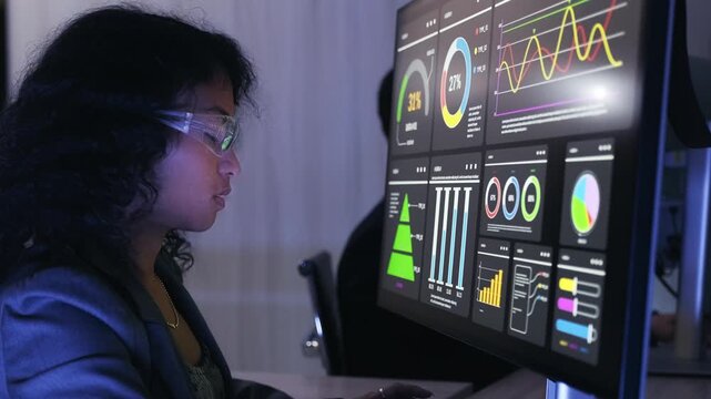 Professional wearing smart glasses analyzes interactive data dashboards on a large monitor in a dim office, reviewing charts, metrics, and trends.