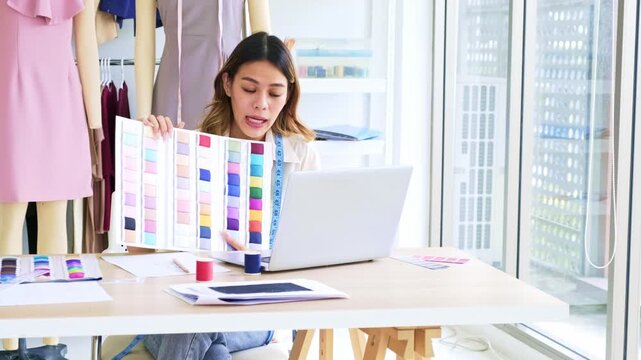 Fashion designer in a bright studio reviews fabric color swatches beside a laptop, thread spools, and measuring tape, planning garment designs with mannequins in the background.