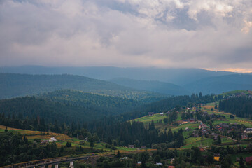 Fototapeta premium Picturesque mountain landscape. Ukrainian Carpathians. A cozy village lies in the valley below, surrounded by rolling green hills and misty mountain peaks. Bright traditional houses and untouched