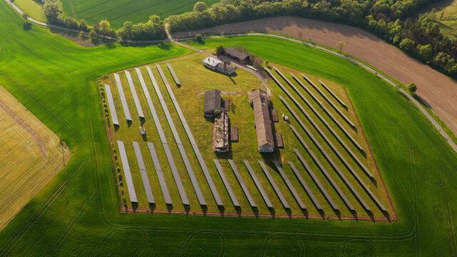 Aerial view of solar panels arranged in rows around farm buildings surrounded by lush green fields and trees during sunset in Chvalkovice, Hradec Kralove Region, Czechia.