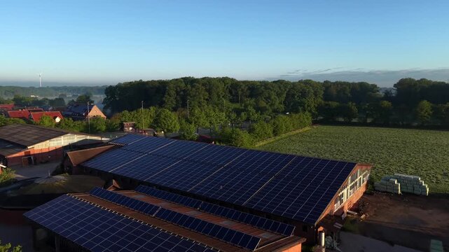 Traditional German farmstead with brick barns and large solar panel arrays on rooftops during misty sunrise over green fields and quiet rural village in Europe during springtime. Aerial flyover shot.