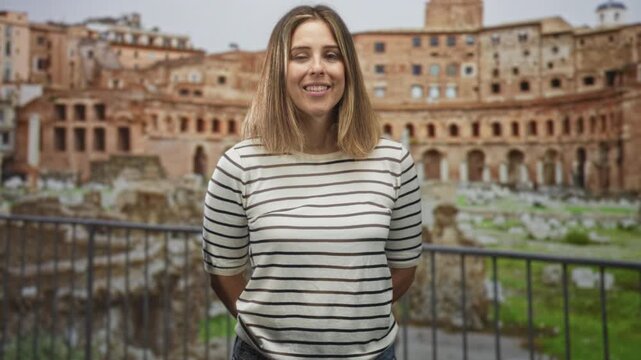 Woman smiling and winking with hands behind back at ancient roman ruins building, wearing striped shirt and denim, nose ring visible; serenity travel.