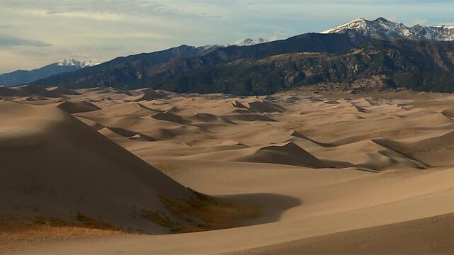 Sweeping sand dunes revealed against rugged snowcapped mountains in Great Sand Dunes National Park, tilt up shot.