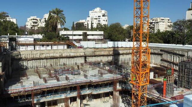 Aerial drone view of a large-scale construction site in Tel Aviv, showing an active tower crane and the structural foundation of a new skyscraper against the city skyline.