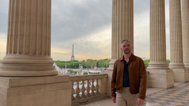 Stylish man walking on a classic colonnaded balcony in Paris, with the iconic Eiffel Tower and the city skyline visible in the background, enjoying a beautiful day during his vacation