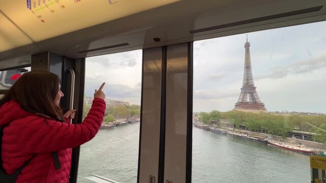 Cheerful young woman enjoying the stunning view of the Eiffel Tower and the Seine River from a train window during her vacation, smiling and pointing at the famous landmark in Paris, France