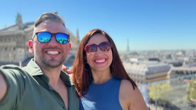 Happy tourist couple taking a selfie video in Paris, France, on a sunny day. The woman kisses the man while they smile and wave with the city skyline and the Eiffel Tower in the background