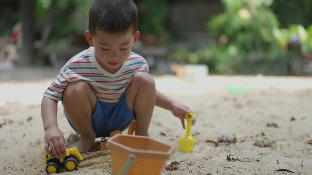 Asian young male child playing sand at beach park, digging with toy truck and bucket, outdoor childhood leisure in natural light, cute boy sandbox fun playground scene