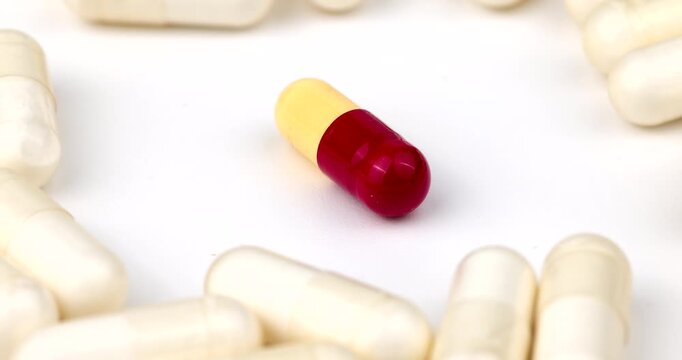 Looping medical video of a bi-color capsule spinning in the center of multiple white pills. Ideal for healthcare, science and drug research use today.