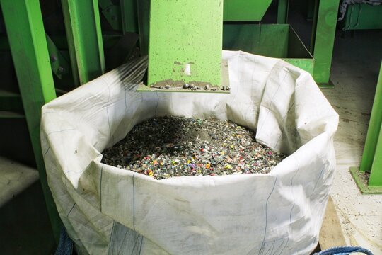 Large white bulk bag filled with shredded electronic waste and plastic scrap material under a green industrial machine hopper dispenser at a recycling plant factory processing facility interior space