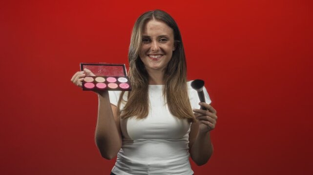 Woman holding makeup palette and brush, smiling and presenting product in studio; playful confidence.