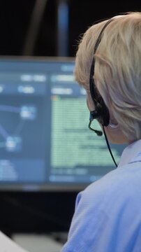 Vertical back view of adult Caucasian coordinator in headset reporting data while working in control center, sitting in front of monitor with programming code