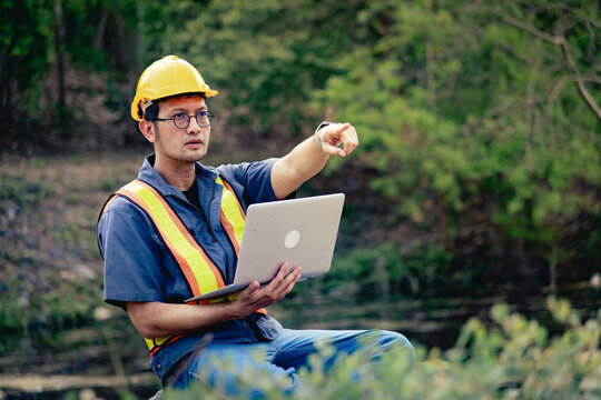 Asian male environmental engineer pointing and checking data alone from laptop, outdoor tree inspection surrounded by forest, nature site evaluation for Environmental Impact Assessment (EIA) 