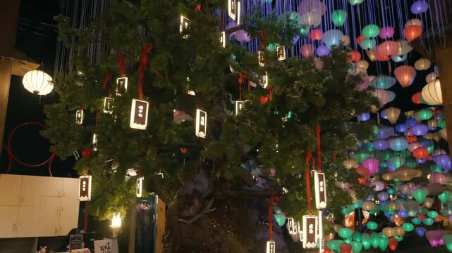 Luminous rectangle shape paper lanterns are hanging on the tree in darkness against the background of magic blue led rain stripes and hundreds of multicolored traditional Vietnames silk lanterns creat