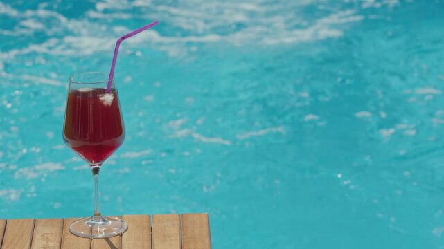 Refreshment glass with red cocktail and straw sitting on wooden table by swimming pool water motion.