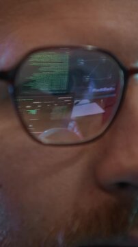 Vertical close up shot of young adult Caucasian network engineer wearing eyeglasses reflecting programming codes while working on computer