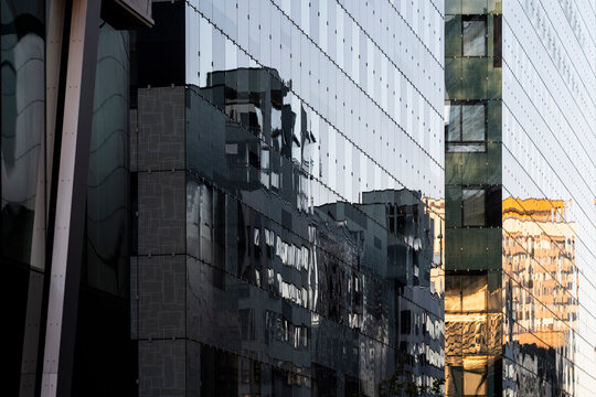 Reflected buildings stretch across glass facade panels, where urban architecture, windows and shadow create layered exterior geometry in daylight