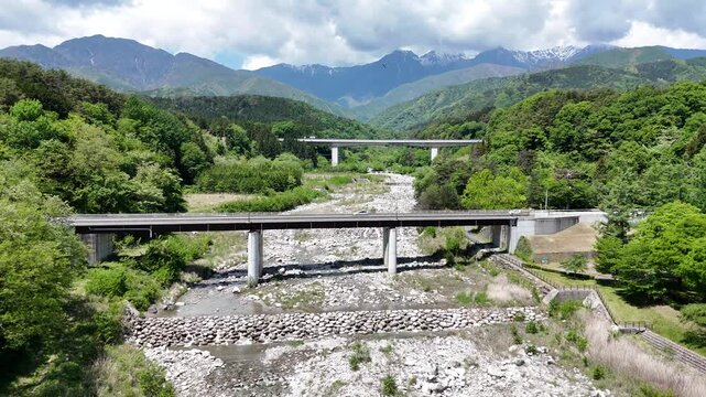 Two bridges span a dry riverbed. Green hills frame distant snow-capped mountains. Rocks and sparse vegetation line the riverbed. Clouds drift above the peaks. No people or animals appear