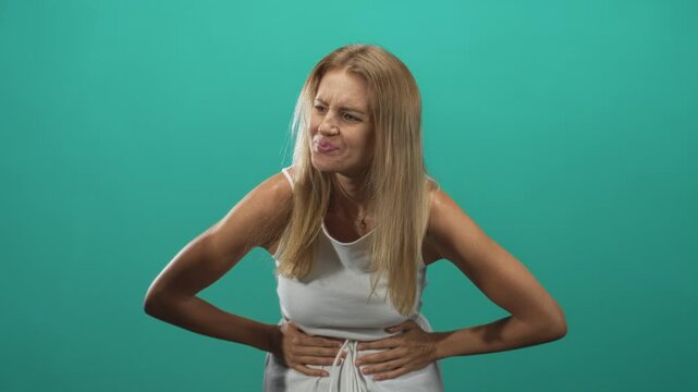 Woman clutching stomach with hands, grimacing and leaning forward in studio wearing white sleeveless dress; discomfort and nausea.