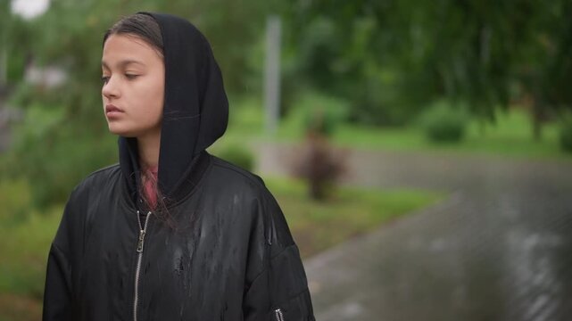 Young woman looking up at sky hooded jacket soaked from drizzle, hand on tree trunk, attentive gaze, soft droplets on hair, park pathway shining, reflective mood, candid lifestyle frame, mindful pause