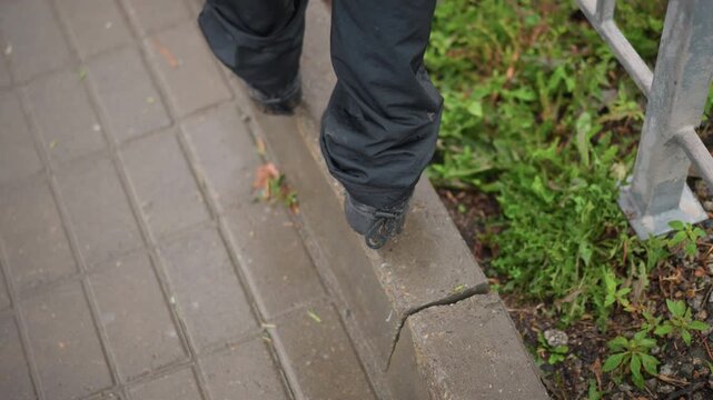 Woman stepping slowly on rain slick curb in quiet mood, closeup of black sneakers and feet on wet brick edge beside green verge and metal railing, contemplative pace, muted color and soft reflections