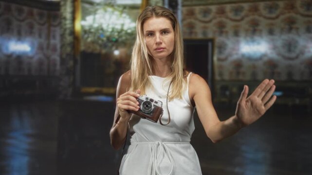 Blonde woman holds vintage camera at chest and raises right hand palm forward in stop gesture inside ornate palace building interior; caution privacy boundary.