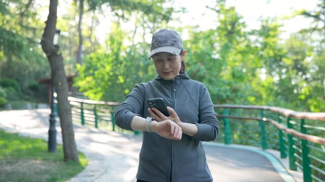 A female adult athlete in casual sportswear checks her smartwatch while exercising on a park paved path surrounded by lush green foliage on a sunny day.