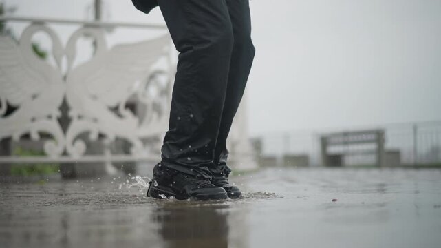 Woman splashing in puddle rainy promenade, black boots kicking water against ornate white railing under overcast sky wet pavement reflects motion and droplets, candid urban mood mixing playful energy