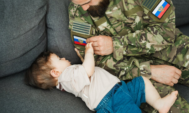 Male russian soldier in military uniform hugs his little son at home while baby boy touching russian flag - Emotional military happy homecoming concept - Main focus on kid hand