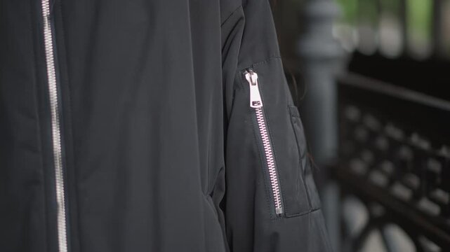 Closeup jacket sleeve zipper detail urban. Rainy outdoor setting, black bomber jacket with textured fabric, silver zipper glinting, hand tucking cuff, subtle motion, commuter preparing for walk,