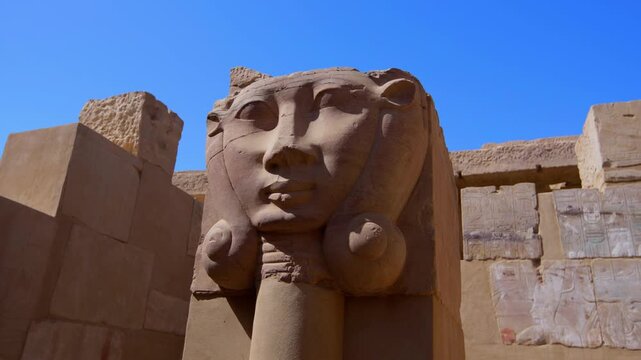 Close-up of ancient Egyptian Hathor-headed column capital with carved goddess face and cow ears on Elephantine Island temple ruins, Aswan, Egypt, against vivid blue sky.