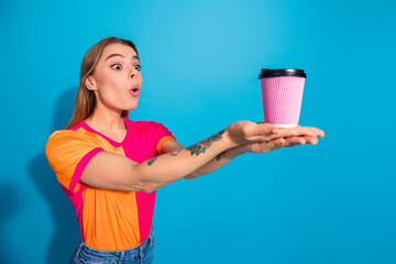 Obraz premium Young woman in bright shirt presents pink coffee cup against blue backdrop with surprised expression