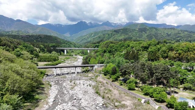 Mountains stretch behind lush green valley. Riverbed flows through stone-filled channel. Concrete bridge spans wide river crossing. Sky clouds drift above forested hills