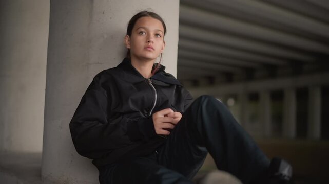 Young woman leaning against concrete pillar, skateboarder resting with gear at feet and phone in hand, raindarkened underpass, soft cinematic light and introspective mood capturing urban respite