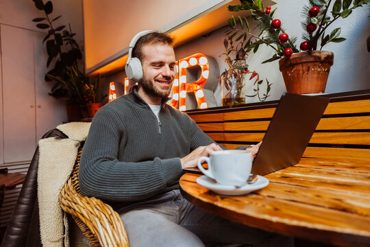 Young adult man smiling while using laptop and wearing headphones in a cafe.