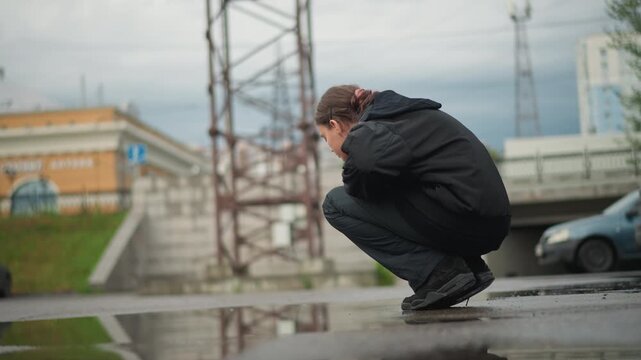 Girl examines water. Young woman tests ripple curiosity. Woman kneeling by reflective puddle exploring ripple. Young lady crouching next to mirrorlike puddle inspecting its surface