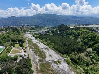 Aerial view of a dry riverbed winding through lush green hills. Clouds drift above distant mountain ridges. Vibrant foliage surrounds the exposed earth. Nature's quiet resilience dominates the scene © Masakazu Tokashiki