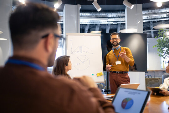 Confident business mentor teaching workshop, presenting data on flipchart to diverse team members in modern office meeting