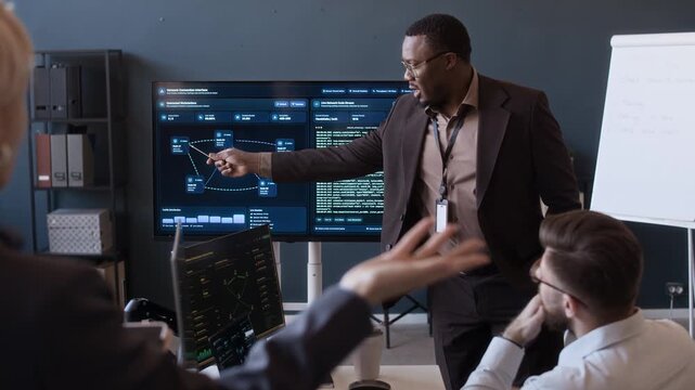 Medium long shot of Caucasian businesspeople listening to Black male network engineer discussing cybersecurity data in modern office